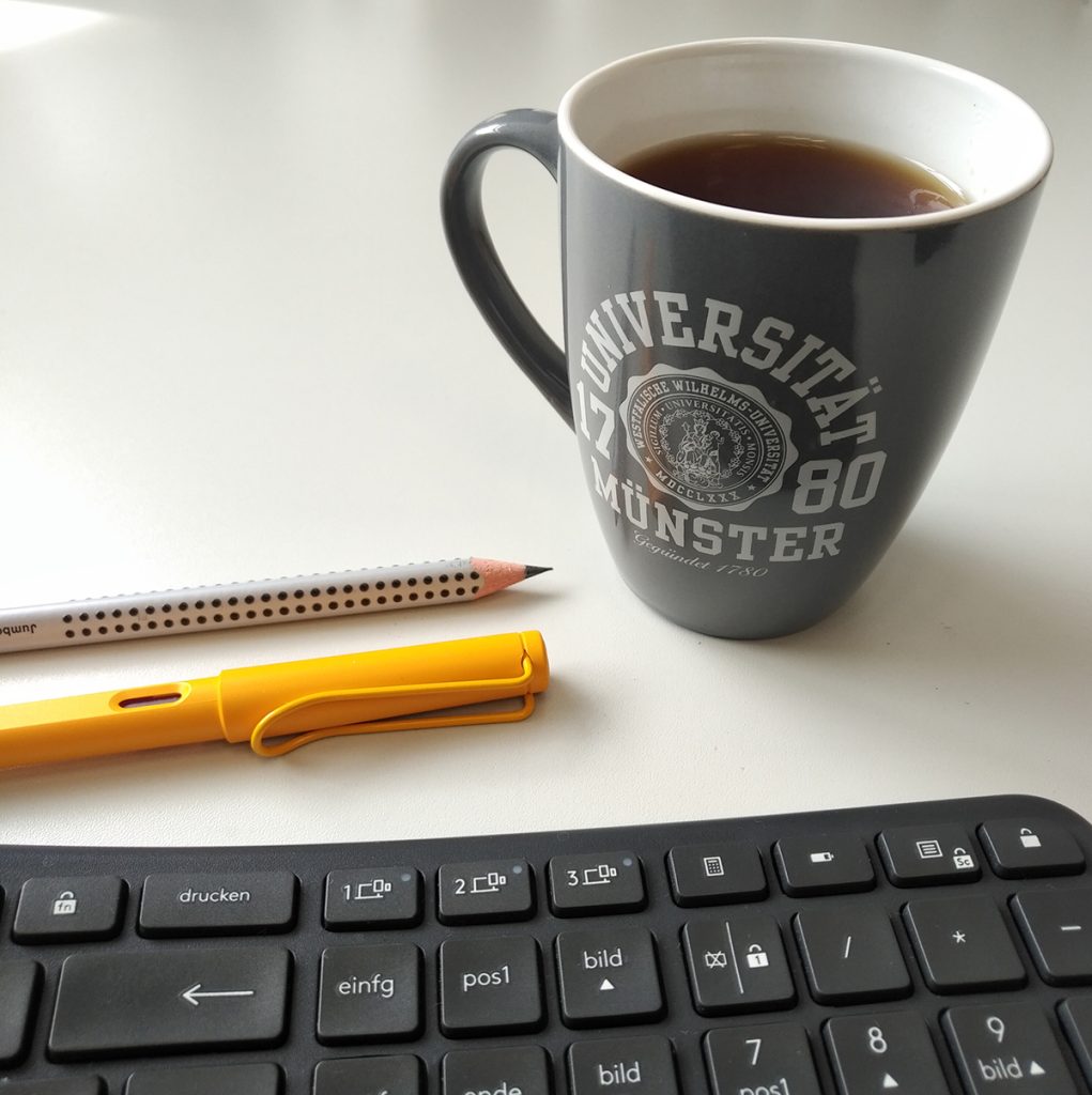 Foto eines Bechers mit der Aufschrift "Universität Münster" hinter einer Tastatur, daneben zwei Stifte